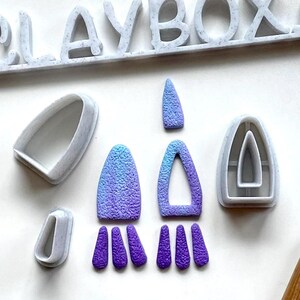 May include: A set of polymer clay earring molds with a gradient blue and purple color scheme. The molds are in the shape of a teardrop, a teardrop with a hole, and a small oval. The molds are on a white surface.