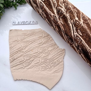 Peut inclure: Un rouleau à pâtisserie en bois marron avec un motif de poissons, à côté d'un morceau d'argile avec le même motif de poissons. Le rouleau à pâtisserie est cylindrique et présente un motif de poissons détaillé gravé.