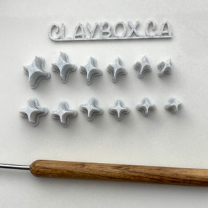 May include: A set of 10 white polymer clay stamps in the shape of a four-pointed star. The stamps are arranged in two rows of five. The stamps are speckled with a light grey colour. The text "CLAYBOXCA" is printed on a white banner above the stamps.