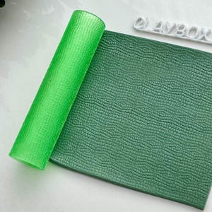 以下が含まれることがあります： 緑のテクスチャ付きの麺棒と緑のテクスチャ付きのポリマークレイのシート。ポリマークレイは、ワニの皮のパターンが施されたテクスチャ付きのシートです。麺棒は透明なプラスチック製で、テクスチャ付きの表面をしています。