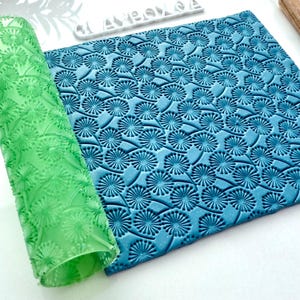 May include: A rolled green textured rolling pin and a blue clay sheet with a repeating floral pattern. The rolling pin is transparent, and the clay sheet is a solid blue color. The pattern is a series of stylized flowers.