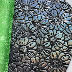 May include: A green rolling pin with a floral design is rolling over a black sheet of fondant with a floral design embossed into it.