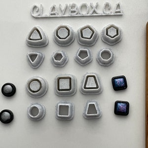 May include: A set of 18 white plastic clay cutters in various shapes, including circles, squares, triangles, and pentagons. The cutters are arranged in rows on a white surface. There are also two small, square, iridescent blue and purple glass tiles.