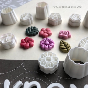 Könnte beinhalten: Ein Set mit 10 weißen Kunststoff-Ausstechern in Form von Blumen und Blättern. Die Ausstecher sind auf einer weißen Oberfläche angeordnet, mit mehreren kleinen, bunten Polymer Clay Blumen und Blättern.