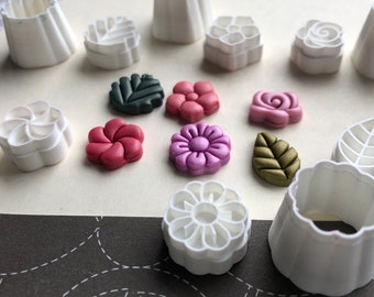 May include: A set of 10 white plastic flower and leaf cookie cutters. The cutters are arranged on a white surface with several small, colorful polymer clay flowers and leaves.