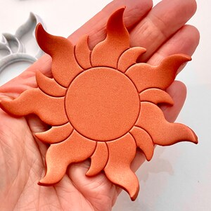 May include: A copper-colored sun with a raised, textured design. The sun has a circular center and 12 points radiating outward.