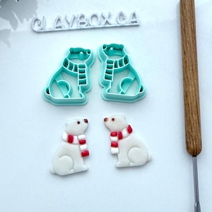 May include: The image displays two polar bear cookie cutters and two finished cookies. The cutters are a light turquoise colour, and the cookies are white with red and white striped scarves. A wooden tool is on the right, and the text "CLAYBOX CA" is at the top.