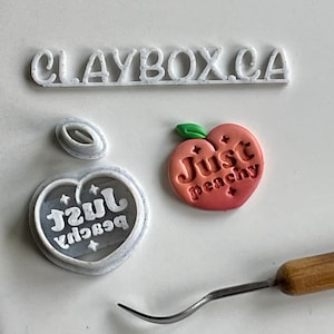 May include: White plastic cookie cutters in the shape of a peach with the text "Just Peachy" inside. The cutters are on a white surface with a wooden tool.