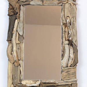 Puede incluir: Espejo rectangular con un marco hecho de varios trozos de madera flotante en tonos marrones y grises. Las piezas de madera flotante están dispuestas para crear una estética rústica y natural. La superficie reflectante del espejo es visible.