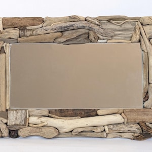 Può includere: Specchio rettangolare con cornice realizzata con vari pezzi di legno trasportato dalle correnti in tonalità marrone, beige e grigio. La superficie riflettente dello specchio è di un colore beige neutro, e i pezzi di legno creano un'estetica rustica e naturale.