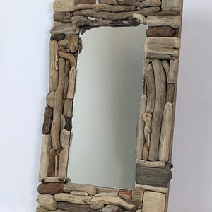 Può includere: Uno specchio rettangolare con una cornice realizzata con vari pezzi di legno trasportato dalle correnti. La cornice è composta da pezzi di legno marrone chiaro, grigio e beige, creando un'estetica naturale e rustica. La superficie riflettente dello specchio è visibile.