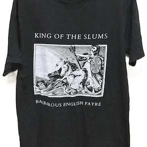 Può includere: T-shirt nero con la scritta "KING OF THE SLUMS" sopra un'illustrazione di una figura con cavalli e la scritta "BARBAROUS ENGLISH FAYRE" sotto. La maglietta è appesa a una gruccia nera.