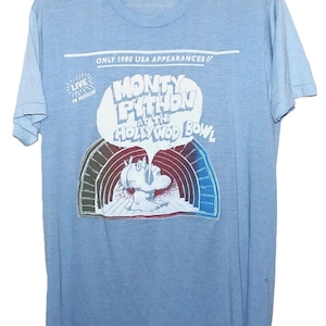 May include: Light blue t-shirt featuring a graphic for the Monty Python at the Hollywood Bowl show. The shirt has the text "ONLY 1980 USA APPEARANCES" and "LIVE IN PERSON". The graphic shows a cartoon image.