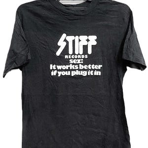 Op de afbeelding: Zwart T-shirt met een witte afbeelding van het woord "STIFF" boven de tekst "RECORDS sez: It works better if you plug it in." Het shirt hangt aan een zwarte hanger.