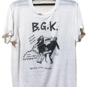 May include: White t-shirt with a black graphic of a person kicking another person, with the text "B.G.K." above the image. The shirt also has the words "sexism, fascism, racism" and "I said stop it now!!".