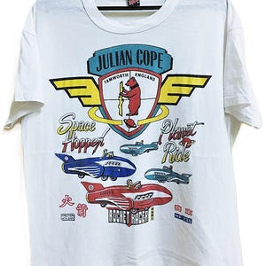 Può includere: T-shirt bianco con un design grafico colorato. Il design include il testo "JULIAN COPE TAMWORTH ENGLAND" sopra un'illustrazione di orso e disegni di razzi retrò con il testo "Space Hopper", "Planet Ride" e "Rocket Racer".