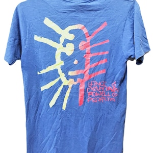 May include: Blue t-shirt with a colorful graphic on the back. The design features a sun-like shape in yellow and red, with text that reads "Lifting Mountain Powell Peralta". The shirt is hanging on a black hanger.
