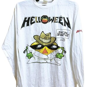 May include: White long-sleeve t-shirt featuring the band name "Helloween" in black block letters. The shirt has a cartoon graphic of a yellow character wearing a hat and sunglasses, holding a newspaper. Text on the shirt reads "I'm not hiding for cover, just can't stand the sun."