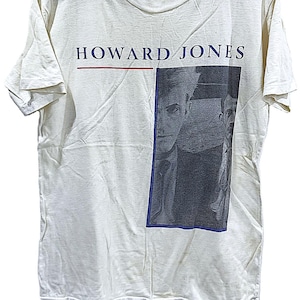 Può includere: T-shirt color crema con il nome "HOWARD JONES" in blu navy sopra una linea rossa. Un'immagine in scala di grigi di una persona è sul lato destro, incorniciata in blu. La maglietta è appesa a una gruccia nera.