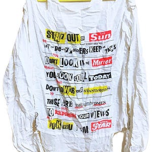 May include: A white long-sleeve shirt featuring distressed cut-out shoulders with metal rings and various printed text in black, red, and yellow. The text includes phrases like STAY OUT OF THE SUN, DON'T LOOK IN THE MIRROR, and FUCK YOU I AM THE STAR.