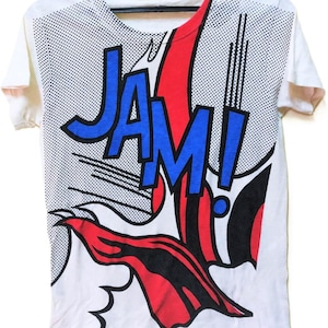 Puede incluir: Camiseta blanca con un gr&aacute;fico estilo c&oacute;mic. El dise&ntilde;o presenta la palabra "JAM!" en letras azules con contornos negros, sobre un estallido rojo y negro. El fondo tiene un patr&oacute;n de puntos.