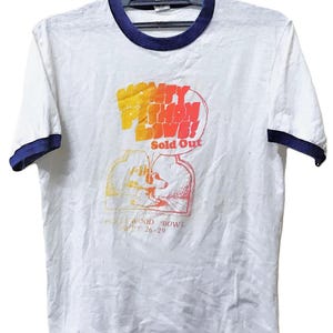 Könnte beinhalten: Weißes Ringer-T-Shirt mit marineblauem Kragen und Ärmelbesatz. Das Shirt zeigt eine Grafik für "Monty Python Live!" im Hollywood Bowl, 26.-29. September. Der Text "Sold Out" ist ebenfalls sichtbar.