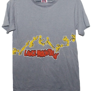 May include: Gray t-shirt featuring a yellow cartoon figure design with the text "LANCE MOUNTAIN" in red and white. The shirt has a crew neck and short sleeves. The shirt is hanging on a black hanger.