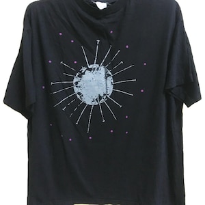 Puede incluir: Camiseta negra con un gráfico gris y blanco de un planeta con líneas radiantes y pequeños puntos morados. La camiseta está colgada en una percha amarilla.