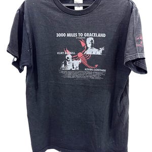 Puede incluir: Una camiseta negra de manga corta con un gráfico de la película 3000 Miles to Graceland. El estampado incluye un escorpión rojo, el texto 3000 Miles to Graceland, Kurt Russell, Kevin Costner y una lista de créditos de reparto y producción.