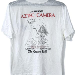 Puede incluir: Camiseta blanca con un gráfico de una mujer de pelo largo y el texto rojo "AZTEC CAMERA". Debajo del gráfico están las palabras "SOLD OUT" y "The Concert Hall, Toronto, March 29, 1985."
