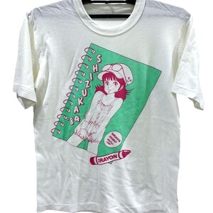 Könnte beinhalten: Ein weißes Kurzarm-T-Shirt mit einer Grafik eines spiralgebundenen Notizbuchs auf grünem Hintergrund. Das Design enthält eine Charakterillustration, den Text SHIZUKA 84, ein kleines kreisförmiges Abzeichen mit YARUKKYA KNIGHT MONTHLY JUMP und einen roten Buntstift.