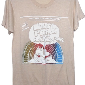 Op de afbeelding: Beige t-shirt met een afbeelding voor de Monty Python at the Hollywood Bowl show. Het ontwerp bevat een regenboogboog en de tekst "ONLY 1980 USA APPEARANCES" en "LIVE IN PERSON".