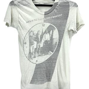 May include: A white short-sleeve t-shirt featuring a gray graphic print of a clock face with three figures inside. The text "DOWN IN THE TUBE STATION AT MIDNIGHT" is printed above the clock graphic. The shirt has a diagonal gray panel design.