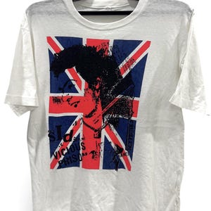 Pode incluir: Uma t-shirt branca de manga curta com uma impressão gráfica da bandeira Union Jack sobreposta por uma silhueta preta de perfil. O texto "Sid Vicious Prison" e "Sex Pistols" está impresso a preto na parte frontal da peça.