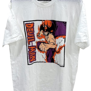 May include: White t-shirt featuring a graphic of a muscular character with orange flames, purple cape, and the text "DEVIL MAN" in orange. The shirt is hanging on a black hanger.
