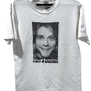 Puede incluir: Una camiseta blanca de manga corta con un estampado gráfico rectangular en blanco y negro de un rostro sonriente. Debajo del retrato, el texto KURT COBAIN está impreso en una fuente negra estilizada. La prenda cuelga de una percha negra.