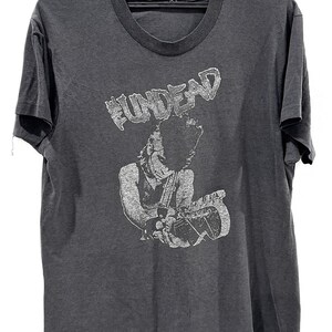 Könnte beinhalten: Ein anthrazitfarbenes Kurzarm-T-Shirt mit einem weißen Grafikdruck einer Person, die eine E-Gitarre spielt. Der Text "FUNDEAD" ist in einer abgenutzten weißen Schriftart über der Illustration aufgedruckt. Das Shirt hat einen Rundhalsausschnitt.