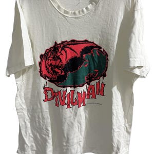 May include: Off-white t-shirt featuring a red and green graphic of a winged demon over a globe, with the word "DEVILMAN" in red flame-style lettering. The shirt is hanging on a black hanger.