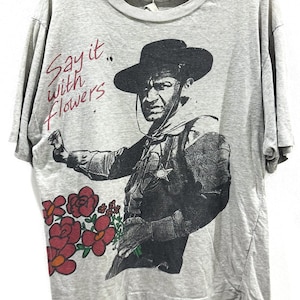 Può includere: T-shirt grigio con grafica vintage di un uomo con cappello che tiene dei fiori, con la scritta "Say it with Flowers". Il design include fiori rossi e rosa. La maglietta ha maniche corte.