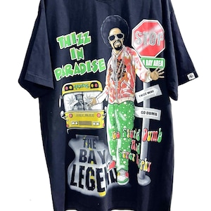 May include: A black short-sleeve t-shirt featuring a graphic print of a person with an afro and sunglasses. The design includes a yellow bus, a red stop sign, and text reading Thizz In Paradise, The Bay Legend, and Go Stupid Dumb And Super Hyphy.