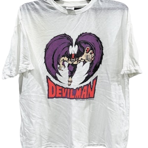 May include: White t-shirt featuring a graphic of Devilman, a character with purple wings and a menacing pose. The word "DEVILMAN" is printed in red, with additional text below. The shirt is hanging on a black hanger.