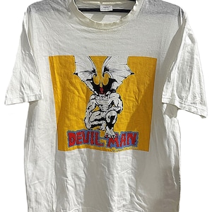 May include: White t-shirt featuring a yellow rectangle with a muscular, winged demon graphic. The text "DEVILMAN" is in red and blue, with "DYNAMIC PLANNING" below. The shirt is hanging on a black hanger.