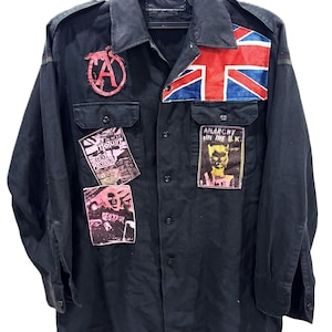 Puede incluir: Una camisa negra de manga larga con botones que presenta un parche rojo con el símbolo de la anarquía, un parche de la bandera Union Jack y dos parches rectangulares con texto que incluye "Anarchy in the U.K.", "Sex Pistols" y "Destroy".