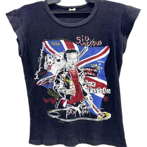 Pode incluir: Uma t-shirt preta sem mangas com uma estampa gráfica de Sid Vicious em frente a uma bandeira Union Jack. O design inclui textos como Sid Vicious, Sex Pistols e Fuck Off, juntamente com ilustrações de uma seringa, um alfinete e correntes.