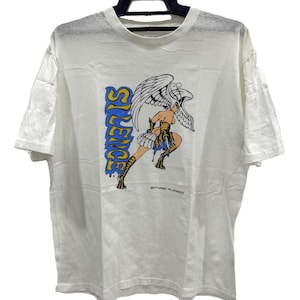 May include: White t-shirt featuring a graphic of a winged figure with the word "SILENCE" in blue and yellow. The figure is in a dynamic pose, with a skirt and boots. The shirt is hanging on a black hanger.