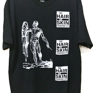 Może przedstawiać: Czarny t-shirt z białą grafiką przedstawiającą postać trzymającą ostrze i draperowaną formę. Koszulka ma trzy białe prostokątne logo z napisem "THE HAIR & SKIN TRADING CO."