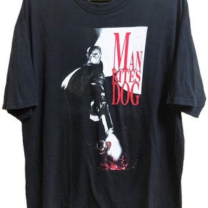 May include: Black t-shirt featuring a graphic from the film "Man Bites Dog." The design includes a black and white image of a person holding a gun, with the film's title in red and white text. The shirt is hanging on a yellow hanger.