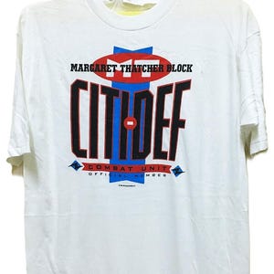 May include: White t-shirt with the text "MARGARET THATCHER BLOCK CITIDEF COMBAT UNIT OFFICIAL MEMBER" in red, blue, and black. The shirt is hanging on a yellow hanger.