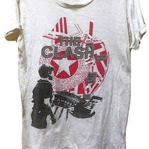 May include: A white short-sleeve t-shirt featuring a graphic print of the band The Clash. The design includes a red star, black text, and a silhouette of a person holding a radio, with various news-style headlines integrated into the circular pattern.