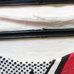 Puede incluir: Primer plano de una camiseta blanca con un estampado de lunares en blanco y negro en la parte inferior izquierda y una raya roja en la parte inferior derecha. La camiseta est&aacute; colgada de una percha negra.